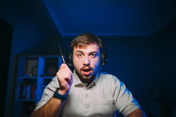 Shocked man in headphones sits at night on the computer and looks at the camera with an emotional face and raises his hand up.