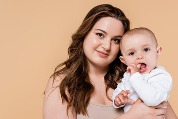 Pretty plus size mother holding baby and looking at camera isolated on beige.