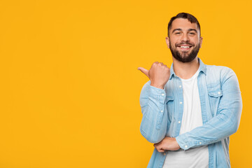 Smiling adult attractive caucasian male points finger towards empty space, isolated on yellow...