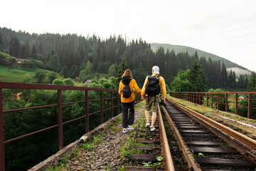 back of a stylish couple of a man and a woman walk on a railway bridge holding hands with backpacks on their backs. Carpathians, Ukraine.