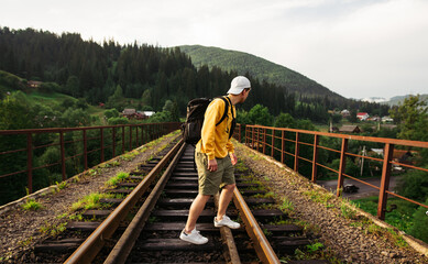 Fototapeta na wymiar Young male tourist stands on a railway viaduct in the mountains against the backdrop of a beautiful landscape and looks to the side, wearing an orange sweatshirt.