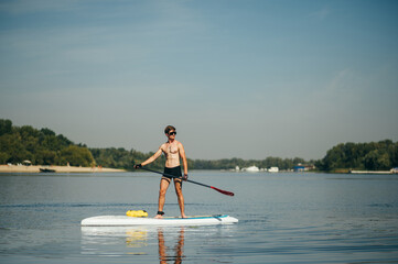 Athletic young man paddles standing on a sup board on the river and looks to the side. A muscular man is active on the water.