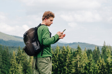 Concentrated tourist with a backpack on a mountain hike uses a smartphone against backdrop of a beautiful landscape with forest and mountains. Guy uses the Internet on a smartphone in the mountains