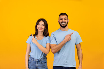 Obraz premium Portrait of excited arab man and woman pointing fingers aside at free space and smiling to camera over yellow background