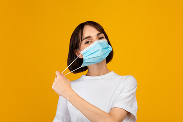 End of the epidemic. Young lady taking off protective face mask, standing isolated over yellow...