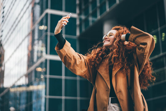 Curly haired young woman wearing fashion clothes with light makeup in glasses makes selfie posing for camera against modern skyscrapers in evening