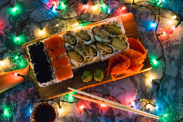 Appetizing rolls with red fish, caviar, mussels, cheese, red ginger, wasabi and soy sauce on a wooden board on a dark background with a christmas garland