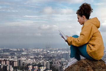 Chica con rizos leyendo libro en montaña con fondo de Barcelona y nubes 