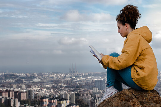 Chica con rizos leyendo libro en monta&ntilde;a con fondo de Barcelona y nubes 