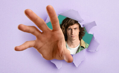 Angry agressive teenage guy reaching through hole in torn violet paper, trying to grab something