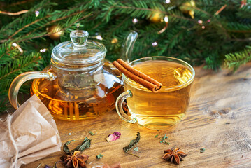 A glass teapot with herbal tea, a cup, a craft bag with herbs on a wooden table with a Christmas tree and garlands
