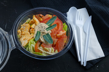 Salad with vegetables and chicken in a plastic container. Takeout food. Salad with cabbage, tomatoes and meat in a disposable bowl with disposable appliances on a black background.