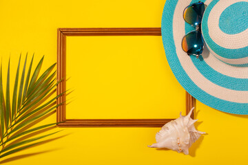 Top view on striped blue hat, sunglasses, palm leaf, seashell and frame for vacation photos on yellow background. Concept of beach holiday, sea tour, warm sunny summer. Copy-paste