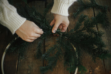 Hands making modern christmas wreath with fir branches on rustic table with round wooden hoop. Atmospheric moody image. Winter holidays preparation. Making minimalistic xmas wreath