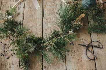 Modern christmas wreath with spruce branches, herbs, scissors, candle on rustic wood. Moody image. Merry Christmas! Winter holidays preparation. Stylish boho xmas wreath