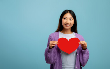 St. Valentines day concept. Playful asian lady holding paper heart and looking aside at free space on blue background