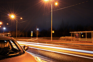 Night city road with parked automobile, street light, bus stop, trails of passing cars and wet asphalt 