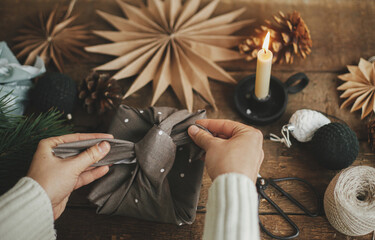 Hands wrapping christmas gift in brown fabric on rustic wooden table with scissors, craft paper...