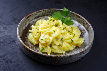 Traditional German potato salad with onion and chives served as close-up in a design bowl on a rustic black board - obrazy, fototapety, plakaty