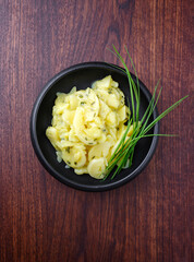 Traditional German potato salad with onion and chives served as top view in a design bowl on a wooden design board