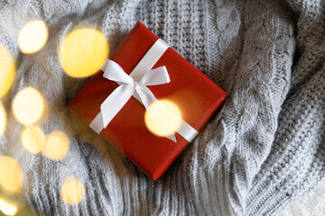 Red kraft gift box with white ribbon on knitted gray sweater with light garland and boke, present for special event, top view. Cozy Christmas still life, birthday or new year, valentines day gift