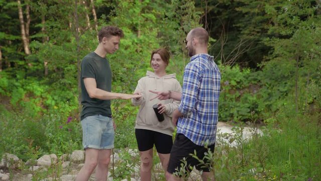 A group of three people playing a rock-paper-scissors game while standing in a forest surrounded by trees on a summer day. Outdoors.