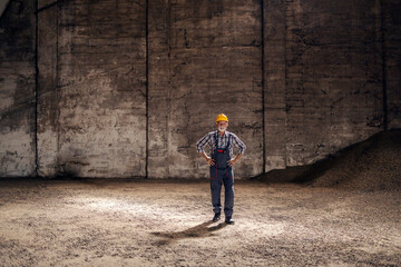 Obraz na płótnie Canvas An old hardworking worker in overalls standing with hands on hips in the dark industrial room full of sugar beet products. Sugar factory, production of sweets