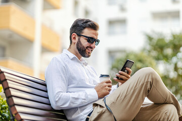 A happy businessman with sunglasses is sitting on a bench in the park, holding coffee to go and using apps on the phone. He holds a dog on a leash.