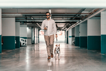 Obraz premium A businessman dressed smart casual, with sunglasses holding coffee to go and walking his dog in the underground garage. A man is searching for his car in the garage. A businessman with a purebred dog