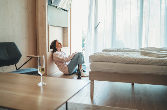 Sad woman sitting on the floor next to window in the bedroom with an opened bottle of alcohol. Unfocused white wine glass on the foreground table. Mental health and alcoholism problems concept image.