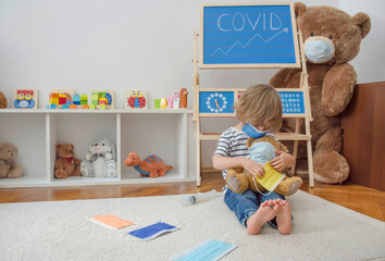 Cute child boy in home quarantine playing doctor on the floor with his sick teddy bears wearing medical masks during coronavirus COVID-2019 and flu outbreak