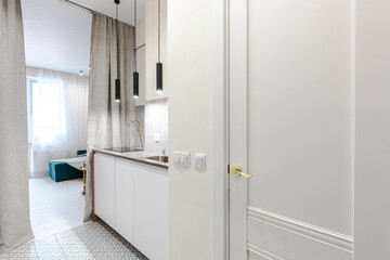 Minimalistic design of a white studio apartment. View from the entrance hall to a compact built-in kitchen, a cozy room with a sofa and a round dining table