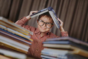 cute caucasian boy wearing glasses covering head with book. Cheerful boy peeking from behind piles...