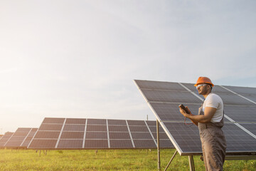 Side view of engineer in orange helmet and brown overalls checking resistance in solar panels outdoors. Indian man using multimeter while working on station.