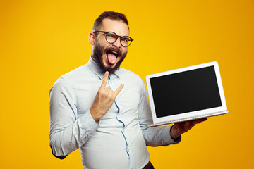 Excited handsome man wearing glasses, showing laptop screen, rock sign and tongue, isolated over yellow background