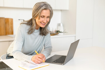 Positive mature woman sitting at the table with laptop in kitchen, studying, researching and taking notes, writing to the notebook, gray-haired senior lady watching online webinar, taking courses