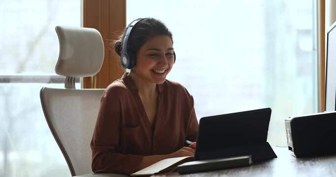 Remote communication. Young Indian woman foreign language teacher wear earphones sit in ergonomic chair by touchpad screen use webcam hold english lesson. Smiling lady expert talk by videocall on pad