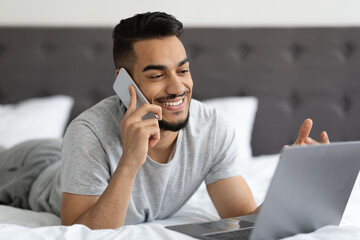 Smiling young arab man talking on cellphone and using laptop in bed
