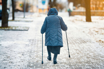 Senior woman with walking canes in hands walks along snowy walkway in winter city street. Old woman...
