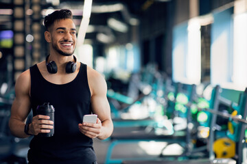 Fototapeta na wymiar Handsome Young Arab Man With Smartphone And Shaker Bottle Posing At Gym
