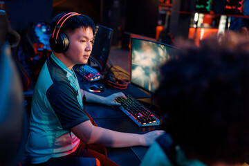 Fototapeta na wymiar Concentrated Asian gamer wearing headphones looking at PC screen of teammate while participating in esports tournament in gaming club