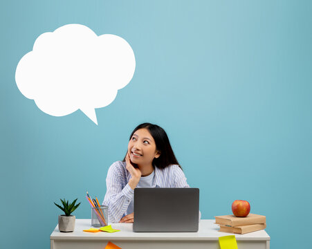 Dreamy asian lady looking up at empty white speech bubble cloud with free space, sitting at desk over blue background