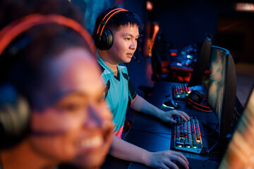 Fototapeta na wymiar Concentrated Asian esports gamer wearing headset looking at PC monitor while practicing in online arena in contemporary e-sports club
