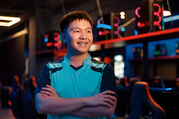 Happy Asian esports player expressing confidence while standing with crossed arms and looking to...