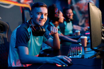 Young man looking at camera and smiling while sitting in gaming chair in interior of dark cyber sports club © Yaroslav Astakhov
