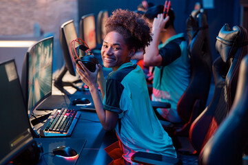 Cheerful African female gamer looking at camera and smiling while holding wired headphones, sitting on gaming chair in gaming club © Yaroslav Astakhov