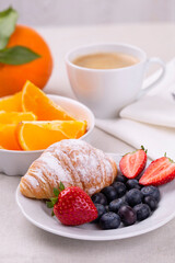 Breakfast with a cup of coffee, fresh croissant, ripe strawberries and blueberries and juicy orange slices on a light table, vertically