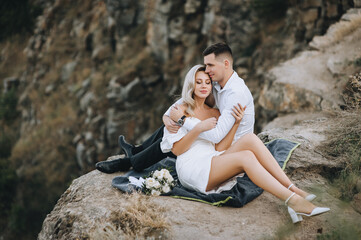 Stylish young groom and a beautiful blonde bride in a white dress are sitting on a rug on a cliff, tenderly embracing, against the backdrop of rocks. Wedding photography.
