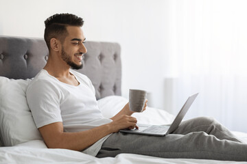 Happy Young Middle Eastern Man Relaxing In Bed With Laptop And Coffee