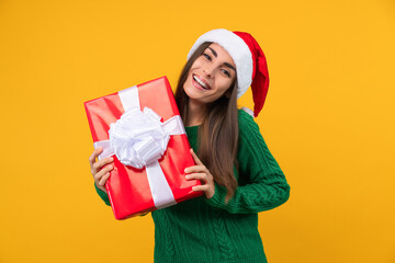 Portrait of smiling pretty girl wearing santa hat and green cozy knitted sweater posing over yellow background with present box in hands